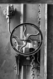 A black and white image shows an industrial metal valve with a circular wheel handle. Rust and wear are visible on the metal surface, and a chain hangs from the mechanism. The background consists of metal panels and rivets.