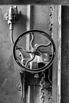 A black and white image shows an industrial metal valve with a circular wheel handle. Rust and wear are visible on the metal surface, and a chain hangs from the mechanism. The background consists of metal panels and rivets.