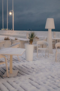 A snowy outdoor scene with patio furniture covered in snow. A potted plant sits in the center amidst the tables and chairs. Large lamp-style lighting fixtures are positioned on the deck, and the area is illuminated by soft lighting against a dark, cloudy sky.