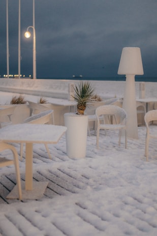 A snowy outdoor scene with patio furniture covered in snow. A potted plant sits in the center amidst the tables and chairs. Large lamp-style lighting fixtures are positioned on the deck, and the area is illuminated by soft lighting against a dark, cloudy sky.