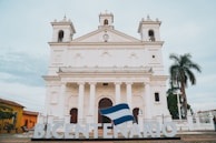 A grand white colonial-style church with two tall bell towers and a central clock is the main focus. The building features large arched windows and is fronted by six columns. There is a large sign in front spelling 'BICENTENARIO,' with a flag draped over the letters. To the right, a tall palm tree and some greenery are visible, while to the left, another building painted in yellow can be seen. The sky above is partly cloudy.