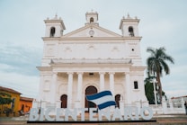 A grand white colonial-style church with two tall bell towers and a central clock is the main focus. The building features large arched windows and is fronted by six columns. There is a large sign in front spelling 'BICENTENARIO,' with a flag draped over the letters. To the right, a tall palm tree and some greenery are visible, while to the left, another building painted in yellow can be seen. The sky above is partly cloudy.