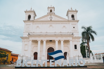 A grand white colonial-style church with two tall bell towers and a central clock is the main focus. The building features large arched windows and is fronted by six columns. There is a large sign in front spelling 'BICENTENARIO,' with a flag draped over the letters. To the right, a tall palm tree and some greenery are visible, while to the left, another building painted in yellow can be seen. The sky above is partly cloudy.