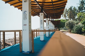 A wooden boardwalk with a series of poles has branding for the Surf City El Salvador ISA World Surfing Games 2021. The boardwalk runs alongside a rocky shoreline, with palm trees and lush greenery on the right side. The structure has an overhanging wooden roof providing shade.