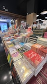 A bookstore display featuring numerous stacks of books arranged on tables. The books vary in size and color, with several covers visible, highlighting a range of titles and authors. The setting is indoors with wooden paneling, and lighting fixtures hang from the ceiling.