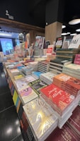 A bookstore display featuring numerous stacks of books arranged on tables. The books vary in size and color, with several covers visible, highlighting a range of titles and authors. The setting is indoors with wooden paneling, and lighting fixtures hang from the ceiling.