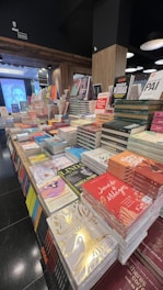 A bookstore display featuring numerous stacks of books arranged on tables. The books vary in size and color, with several covers visible, highlighting a range of titles and authors. The setting is indoors with wooden paneling, and lighting fixtures hang from the ceiling.