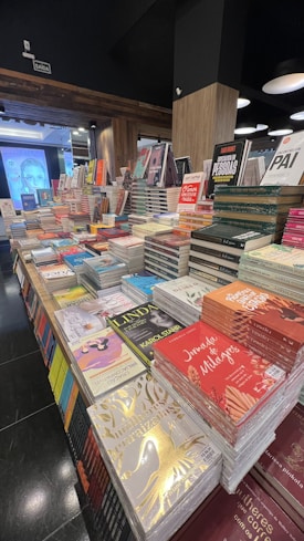 A bookstore display featuring numerous stacks of books arranged on tables. The books vary in size and color, with several covers visible, highlighting a range of titles and authors. The setting is indoors with wooden paneling, and lighting fixtures hang from the ceiling.