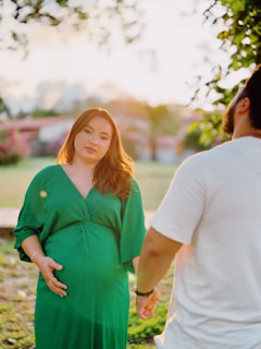 A pregnant woman in a green dress stands holding hands with a man wearing a white shirt. They are outdoors, possibly in a park or garden, with sunlight creating a warm and serene atmosphere.