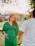 A pregnant woman in a green dress stands holding hands with a man wearing a white shirt. They are outdoors, possibly in a park or garden, with sunlight creating a warm and serene atmosphere.