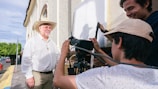Three people are engaged in a filming activity outdoors. One person, wearing a cowboy hat and a cross necklace, stands in front of the camera. Two others appear to be filming, with one operating a camera and equipment. The background features a building and a street lined with trees.