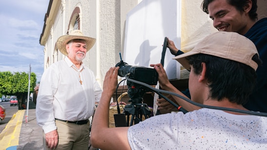 Three people are engaged in a filming activity outdoors. One person, wearing a cowboy hat and a cross necklace, stands in front of the camera. Two others appear to be filming, with one operating a camera and equipment. The background features a building and a street lined with trees.