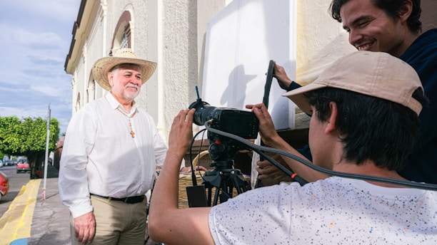 Three people are engaged in a filming activity outdoors. One person, wearing a cowboy hat and a cross necklace, stands in front of the camera. Two others appear to be filming, with one operating a camera and equipment. The background features a building and a street lined with trees.