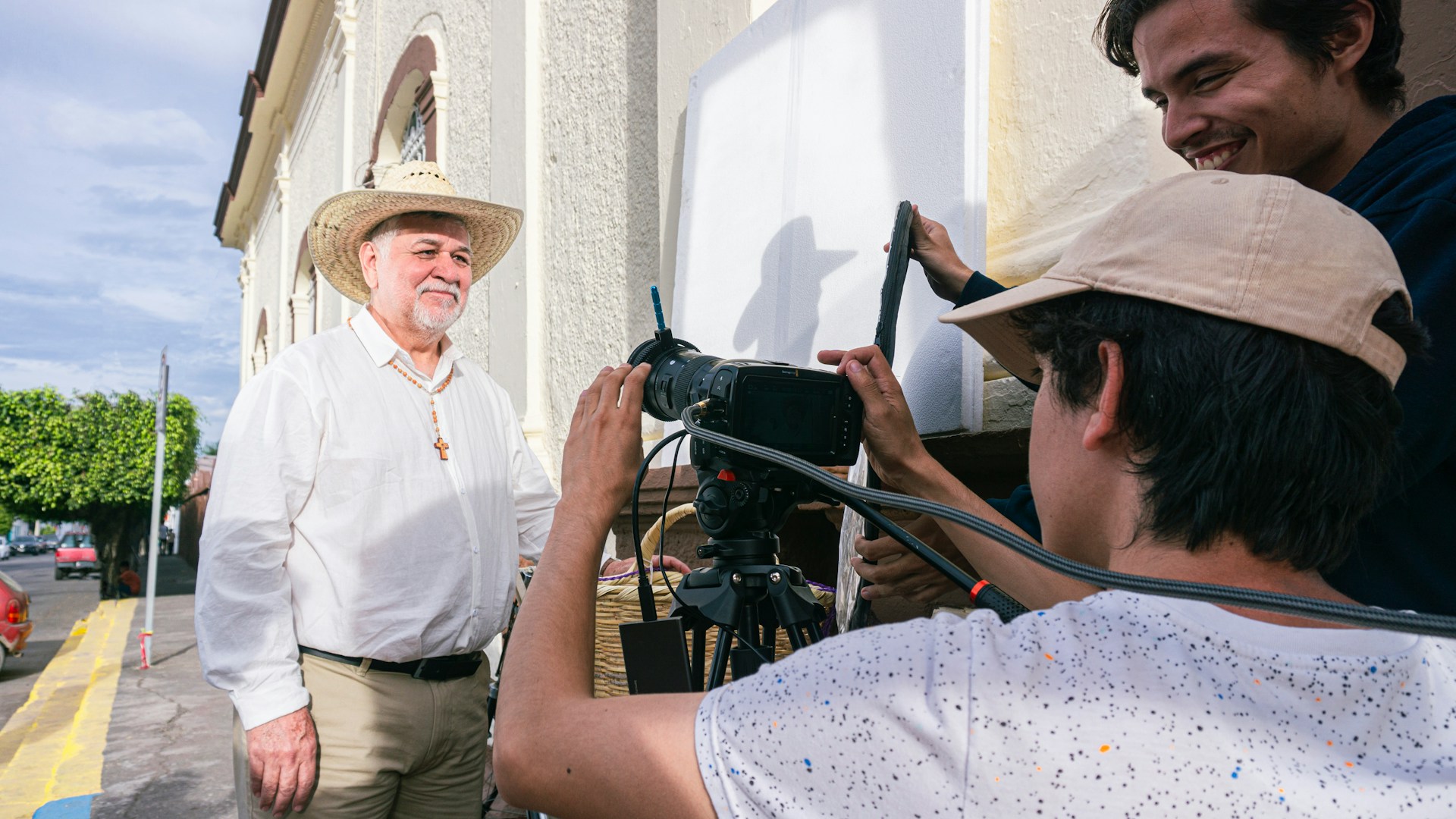 Three people are engaged in a filming activity outdoors. One person, wearing a cowboy hat and a cross necklace, stands in front of the camera. Two others appear to be filming, with one operating a camera and equipment. The background features a building and a street lined with trees.