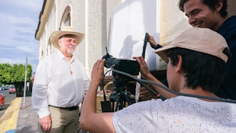Three people are engaged in a filming activity outdoors. One person, wearing a cowboy hat and a cross necklace, stands in front of the camera. Two others appear to be filming, with one operating a camera and equipment. The background features a building and a street lined with trees.