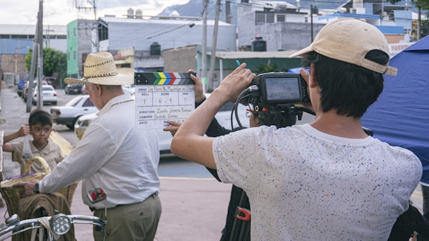 A film crew captures a scene on an urban street. One person holds a clapperboard, while another operates a camera. A man wearing a straw hat and carrying a phone stands in the foreground. A child with a basket on a bicycle observes the scene. Buildings and parked cars line the background.