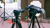 A professional camera setup on a tripod is positioned outdoors with various cables and audio equipment visible. The camera is pointed towards an area with a white gate in the background and some red and black wiring running along the ground. Next to the camera is a field audio recorder mounted on another tripod, covered with a protective case.
