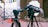 A professional camera setup on a tripod is positioned outdoors with various cables and audio equipment visible. The camera is pointed towards an area with a white gate in the background and some red and black wiring running along the ground. Next to the camera is a field audio recorder mounted on another tripod, covered with a protective case.