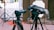 A professional camera setup on a tripod is positioned outdoors with various cables and audio equipment visible. The camera is pointed towards an area with a white gate in the background and some red and black wiring running along the ground. Next to the camera is a field audio recorder mounted on another tripod, covered with a protective case.