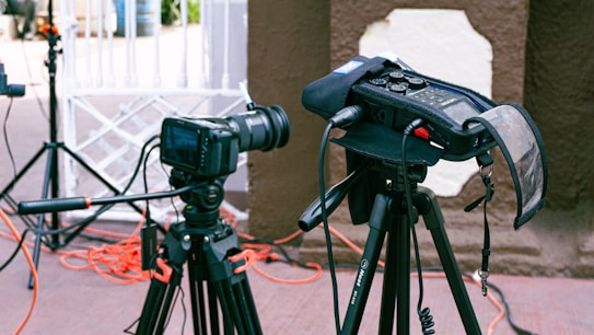 A professional camera setup on a tripod is positioned outdoors with various cables and audio equipment visible. The camera is pointed towards an area with a white gate in the background and some red and black wiring running along the ground. Next to the camera is a field audio recorder mounted on another tripod, covered with a protective case.