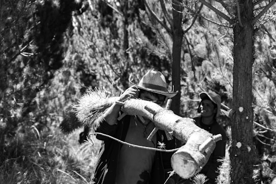 A black and white photograph of two people in a forested area, where one of them is carrying a large tree branch on their shoulder. The person at the forefront is wearing a hat and is partially obscured by the branch. Trees and foliage are surrounding them, creating a dense, natural environment.