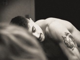 A black and white image of a shirtless person with short hair and a tattoo of a wolf on their upper arm. The tattoo appears detailed, and their expression seems contemplative. Another blurred figure is present in the foreground, creating a sense of depth.
