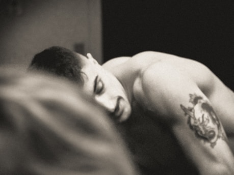 A black and white image of a shirtless person with short hair and a tattoo of a wolf on their upper arm. The tattoo appears detailed, and their expression seems contemplative. Another blurred figure is present in the foreground, creating a sense of depth.