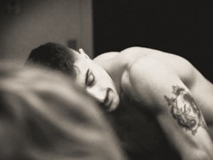A black and white image of a shirtless person with short hair and a tattoo of a wolf on their upper arm. The tattoo appears detailed, and their expression seems contemplative. Another blurred figure is present in the foreground, creating a sense of depth.