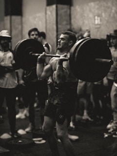 A person is engaged in a weightlifting exercise, performing a barbell lift in a gym environment. The focus is on the concentration and effort of the lifter, while people watch in the background.