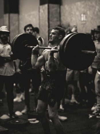 A person is engaged in a weightlifting exercise, performing a barbell lift in a gym environment. The focus is on the concentration and effort of the lifter, while people watch in the background.