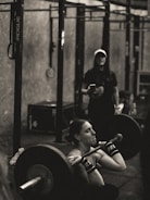 A person is intensely focused while performing a barbell squat in a gym environment. The atmosphere appears rugged with metal gym equipment, and another individual stands in the background observing or coaching.
