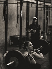 A person is intensely focused while performing a barbell squat in a gym environment. The atmosphere appears rugged with metal gym equipment, and another individual stands in the background observing or coaching.