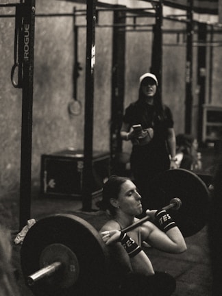 A person is intensely focused while performing a barbell squat in a gym environment. The atmosphere appears rugged with metal gym equipment, and another individual stands in the background observing or coaching.
