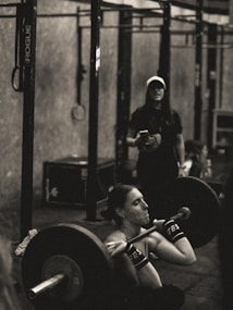 A person is intensely focused while performing a barbell squat in a gym environment. The atmosphere appears rugged with metal gym equipment, and another individual stands in the background observing or coaching.