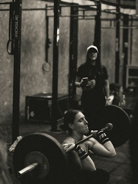 A person is intensely focused while performing a barbell squat in a gym environment. The atmosphere appears rugged with metal gym equipment, and another individual stands in the background observing or coaching.