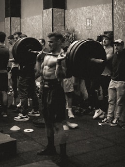 A black and white photograph of a muscular person performing a squat with a heavily loaded barbell across their shoulders, surrounded by spectators in a gym setting. The image conveys a sense of strength and focus.