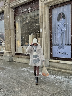 A person wearing a thick white fur coat, a white beanie, and a scarf walks past a store named 'La Mania' during light snowfall. The person is also carrying a netted bag with items that have orange colors. There is a large advertisement poster of a woman in a grey suit and sunglasses in the background.