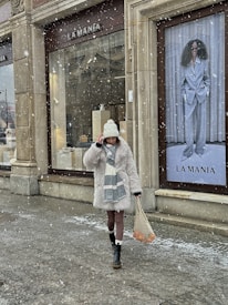 A person wearing a thick white fur coat, a white beanie, and a scarf walks past a store named 'La Mania' during light snowfall. The person is also carrying a netted bag with items that have orange colors. There is a large advertisement poster of a woman in a grey suit and sunglasses in the background.
