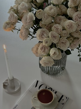 A bouquet of pale pink roses in a ribbed glass vase is placed on a white surface. Next to the vase is an elegant white candle in a simple holder, with the flame gently flickering. A stylish teacup filled with coffee sits on a magazine, which is partially visible with the title 'Vogue'. The scene is minimalistic and sophisticated, exuding a calm and serene atmosphere.