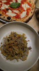 A plate of pasta topped with a creamy sauce and sprinkled with crushed nuts is placed alongside a classic Margherita pizza with a fresh basil leaf at the center. The crust of the pizza is golden and slightly charred, with melted cheese and vibrant red tomato sauce visible.