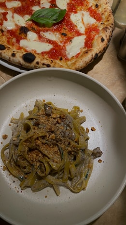A plate of pasta topped with a creamy sauce and sprinkled with crushed nuts is placed alongside a classic Margherita pizza with a fresh basil leaf at the center. The crust of the pizza is golden and slightly charred, with melted cheese and vibrant red tomato sauce visible.