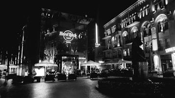 A black and white nighttime scene of an urban area featuring a Hard Rock Cafe with illuminated signs and outdoor seating. Nearby buildings showcase elaborate architectural details and are also brightly lit. There are several umbrellas and tables with people sitting and possibly enjoying the night. The scene has an overall bustling and lively atmosphere.