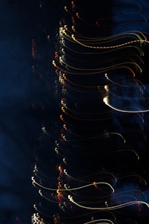 A dark image features a series of curving, colorful light streaks. These streaks are predominantly white, yellow, and red and appear to be captured using long exposure or slow shutter speed techniques, creating an abstract, wavy effect.