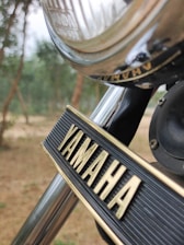 A close-up view of a motorcycle with the brand name Yamaha prominently displayed on a decorative metal plate. The shiny chrome surface and clean lines indicate a well-maintained piece of machinery. The background shows a blurred outdoor setting with trees.