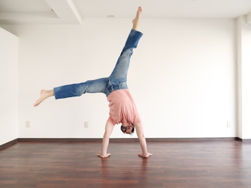 A person performing a handstand in a minimalist, bright room with wooden flooring. The person is wearing a pink top and blue jeans, and the space is largely empty, emphasizing the act of balancing.