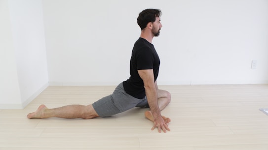 A person is performing a yoga pose resembling the pigeon pose on the floor. The person is positioned with their front leg bent at the knee while the back leg is extended straight back. They are wearing a black t-shirt and gray shorts, and they are placed in a minimalistic room with light-colored wooden flooring and white walls.