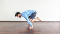 A person in a blue long-sleeve T-shirt and dark shorts is performing a yoga or fitness pose on a wooden floor. They are balancing on their hands with their legs bent and lifted off the ground. The room is minimally decorated with a white wall in the background.