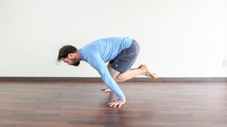 A person in a blue long-sleeve T-shirt and dark shorts is performing a yoga or fitness pose on a wooden floor. They are balancing on their hands with their legs bent and lifted off the ground. The room is minimally decorated with a white wall in the background.