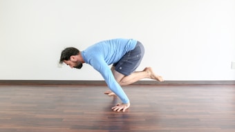 A person in a blue long-sleeve T-shirt and dark shorts is performing a yoga or fitness pose on a wooden floor. They are balancing on their hands with their legs bent and lifted off the ground. The room is minimally decorated with a white wall in the background.