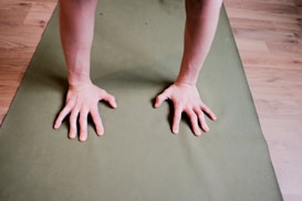 Two hands are placed on a green yoga mat, presumably in a position for a yoga pose or exercise. The floor is wooden with a light brown finish.